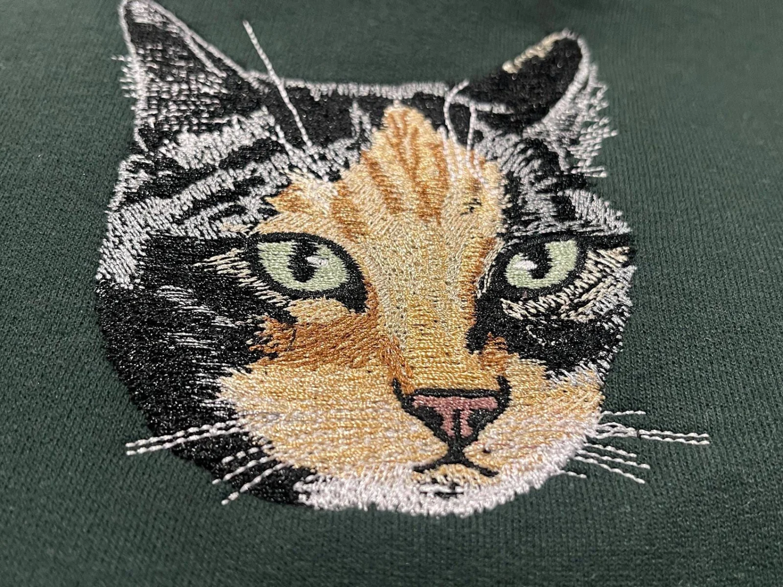Close-up of a thread-painted calico cat face with green eyes embroidered on a forest green sweatshirt