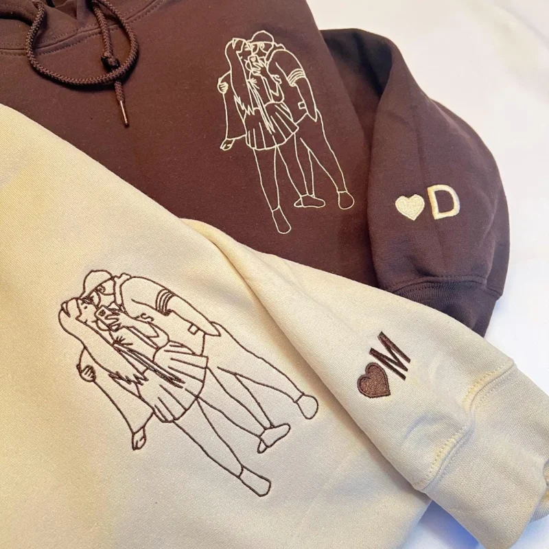 Dark brown and sand hoodies with single-line couple portraits on the chest and a heart with D or M on the sleeves