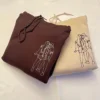 Folded dark brown and sand hoodies showing the embroidered line-art couple in cream and brown thread