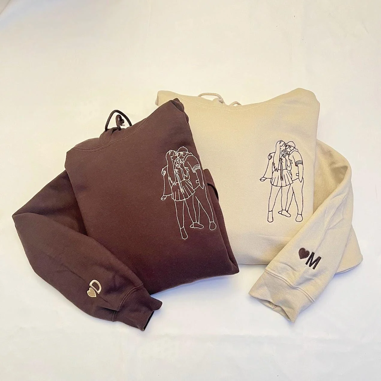 Matching dark brown and sand hoodies laid flat, each with an outline couple portrait and a heart initial cuff