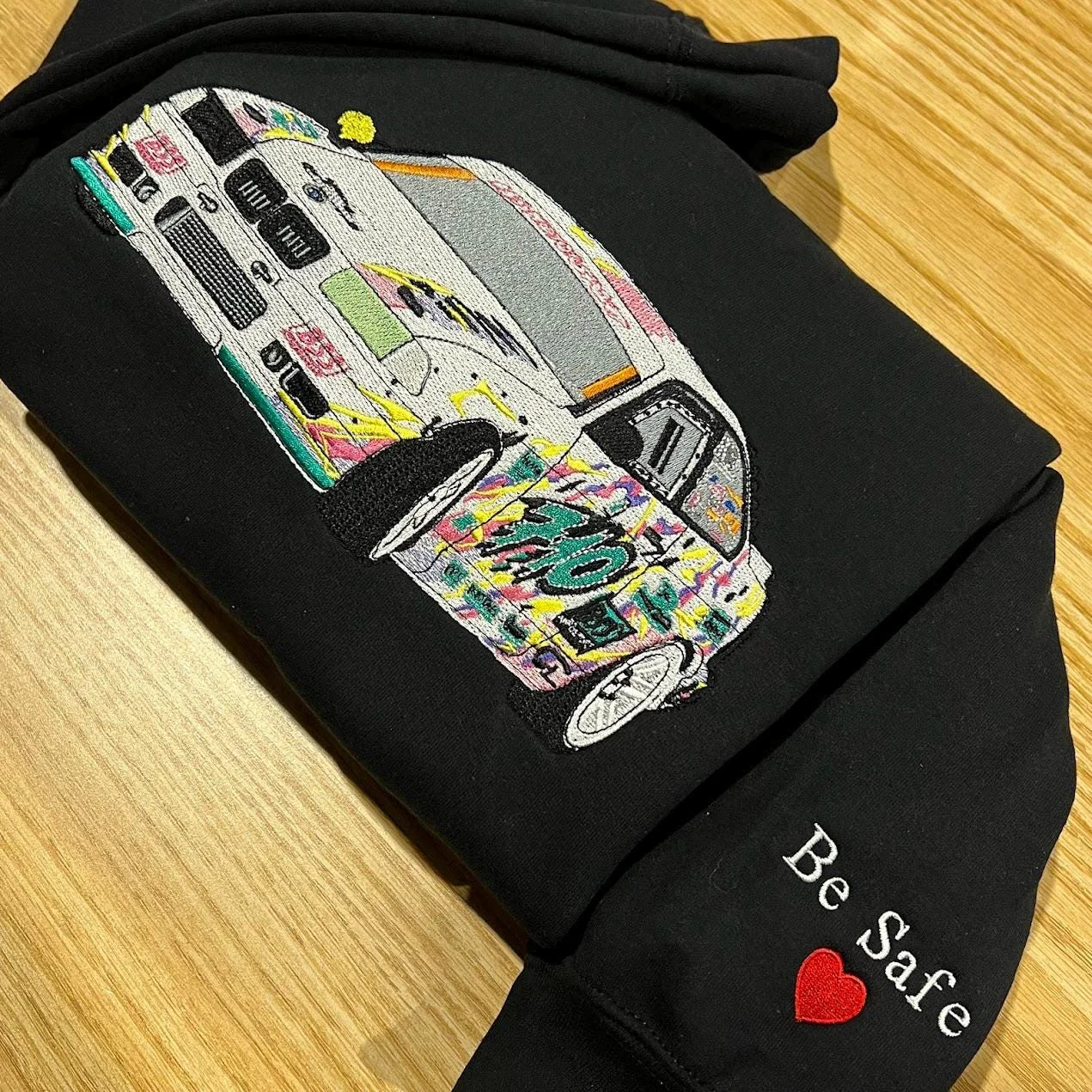 Black hoodie on a wooden table with the graffiti sports car on the chest and Be Safe with a red heart on the cuff