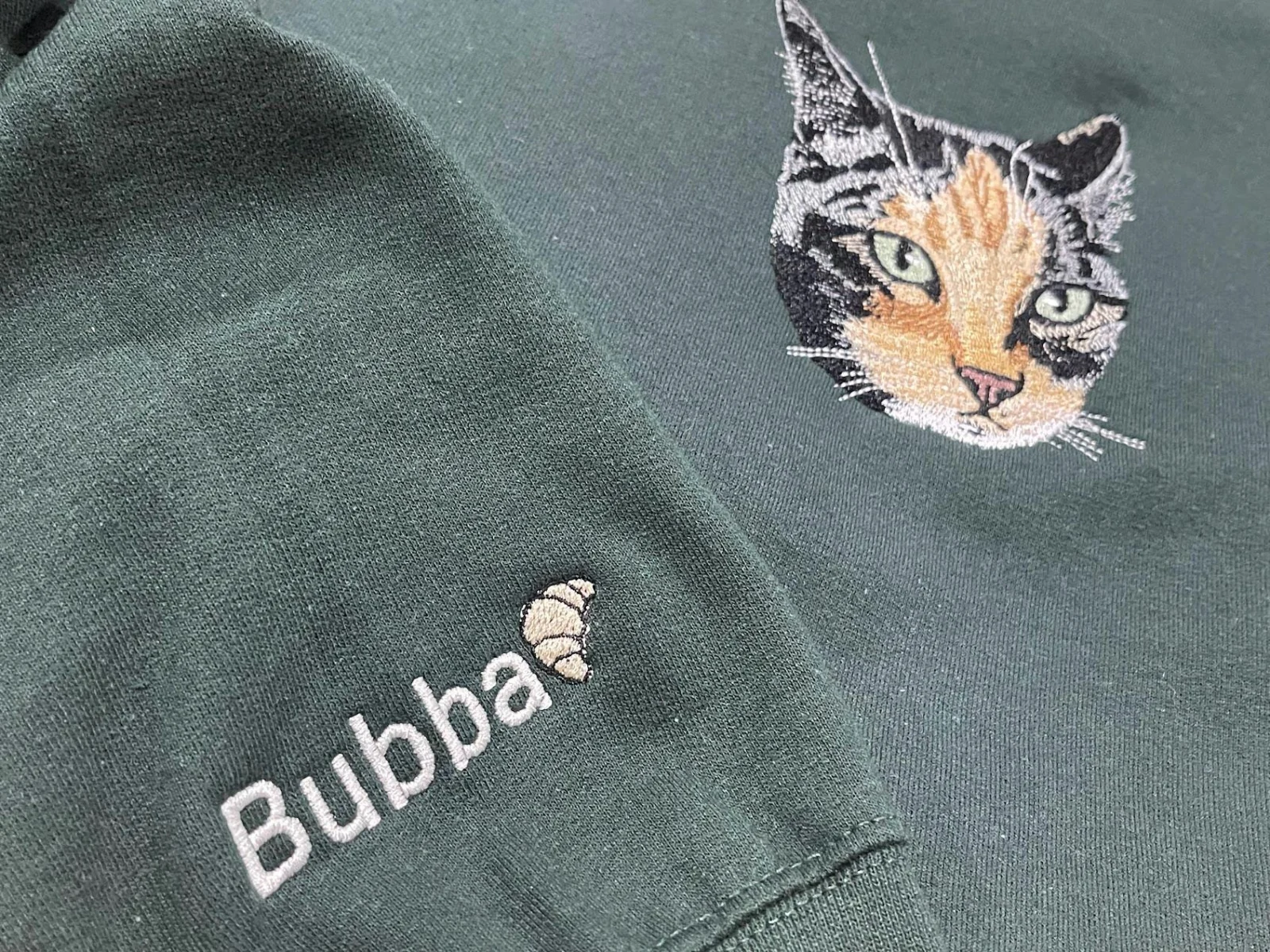 Forest green sweatshirt with an embroidered calico cat face on the chest and the name Bubba with a croissant on the sleeve