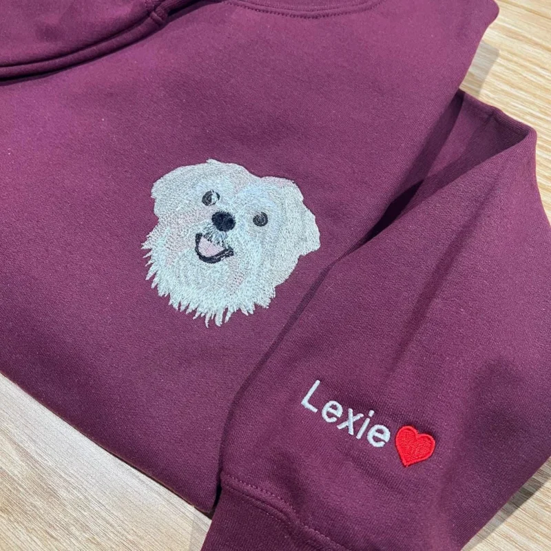 Maroon sweatshirt with an embroidered white fluffy dog face and the name Lexie with a red heart on the sleeve