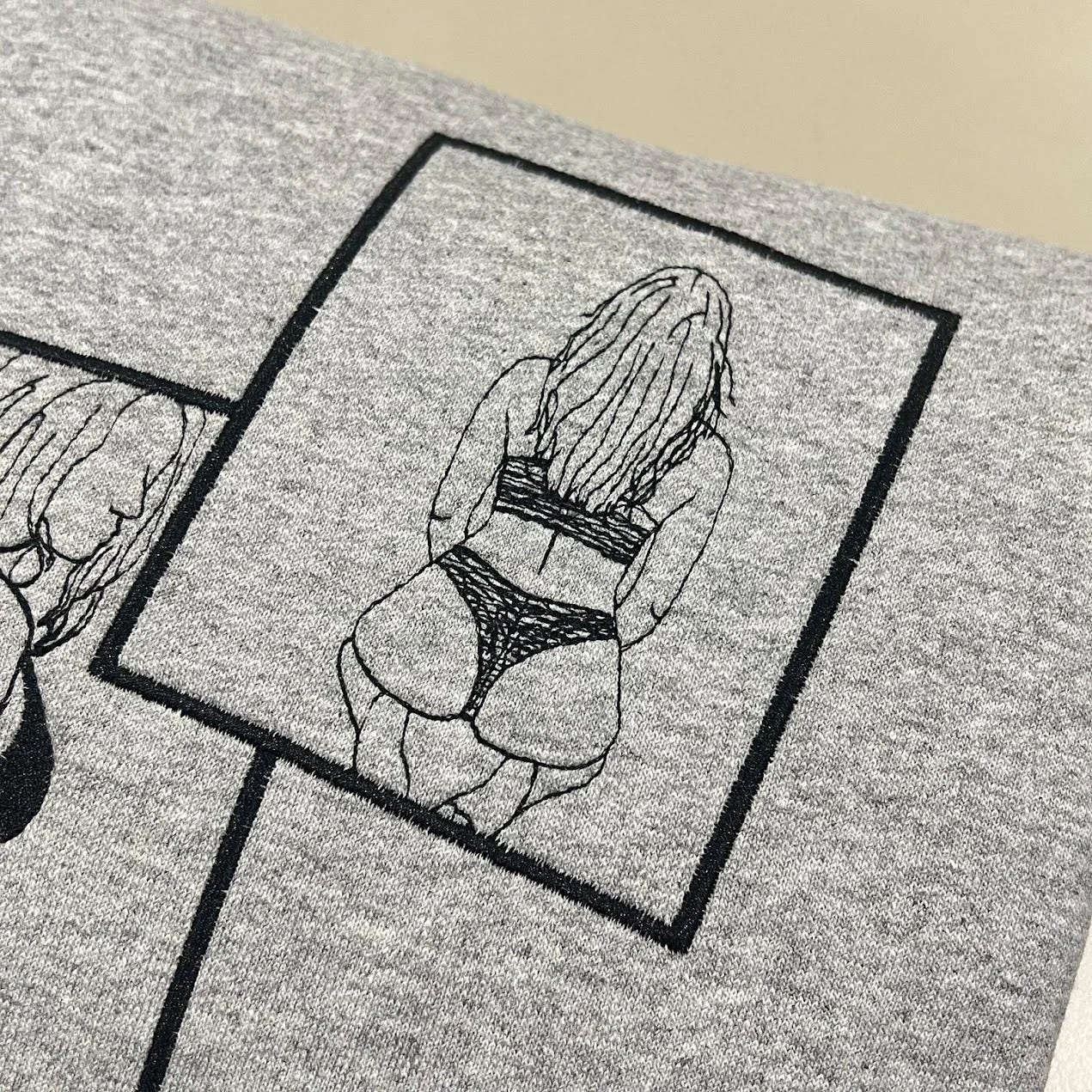 Close-up of a framed black outline drawing of a seated woman in a bikini, seen from behind, on grey fleece