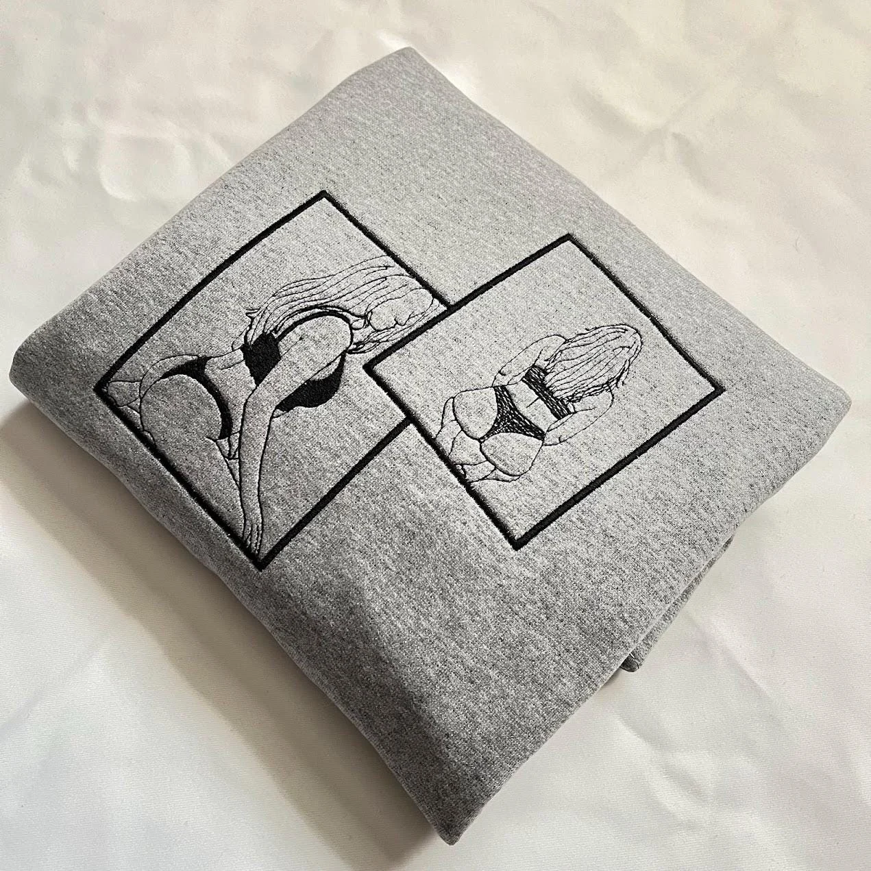 Grey sweatshirt folded at an angle with two framed outline portraits embroidered in black thread
