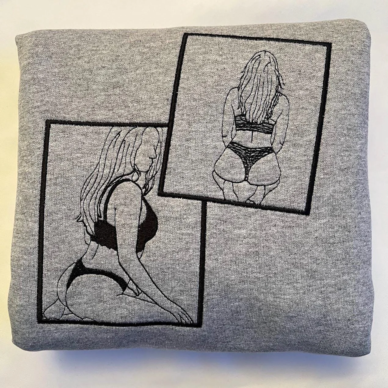 Folded grey sweatshirt with two overlapping framed black line-art figures stitched across the front