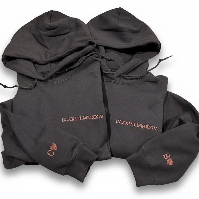 Two dark grey hoodies with IX.XXVII.MMXXIV in rose-gold Roman numerals on the chest and C or S with a heart on the cuffs