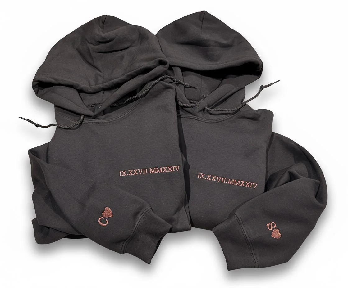 Two dark grey hoodies with IX.XXVII.MMXXIV in rose-gold Roman numerals on the chest and C or S with a heart on the cuffs