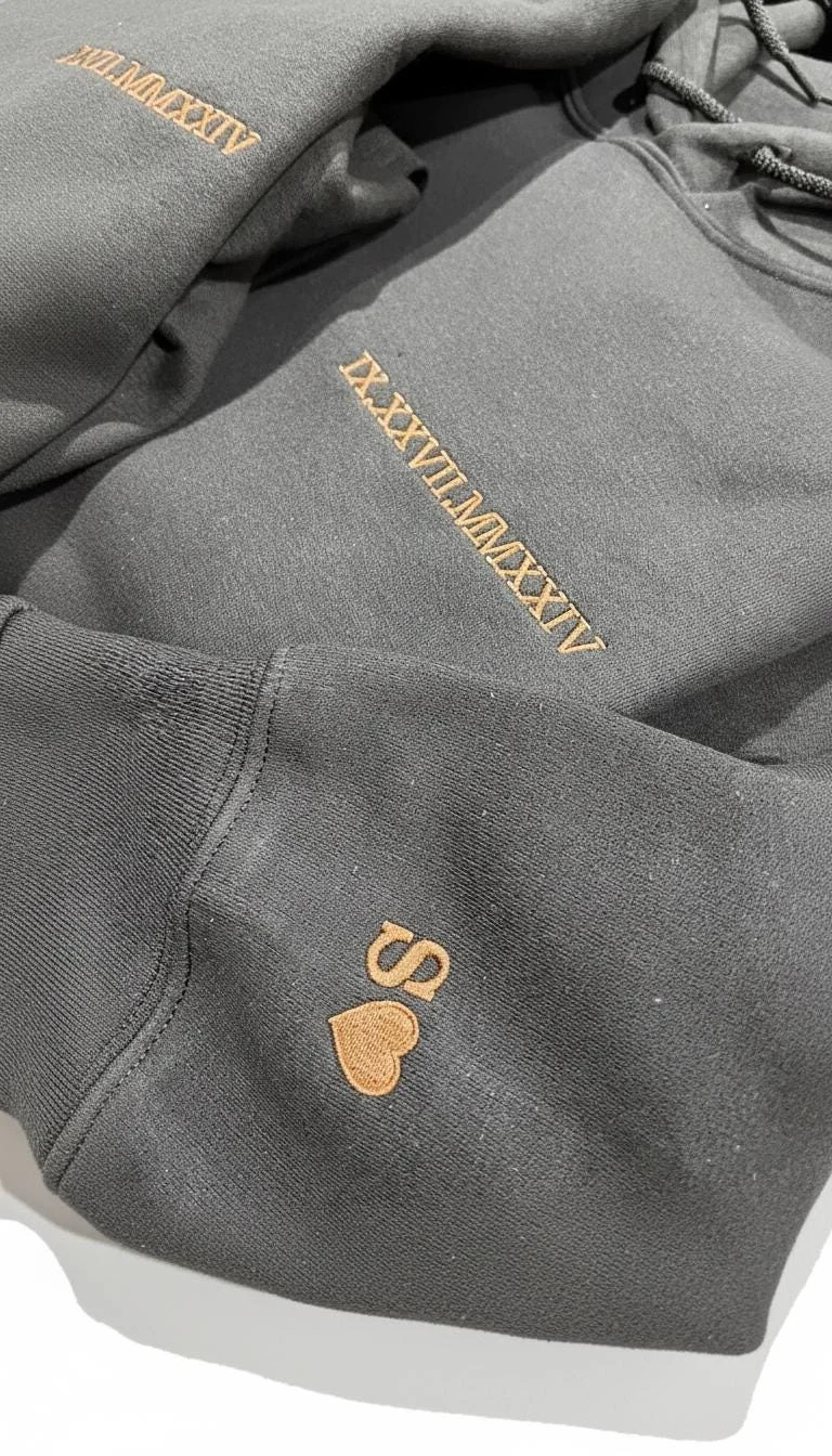 Dark grey hoodie with the date IX.XXVII.MMXXIV stitched in copper thread and an S with a heart on the sleeve cuff