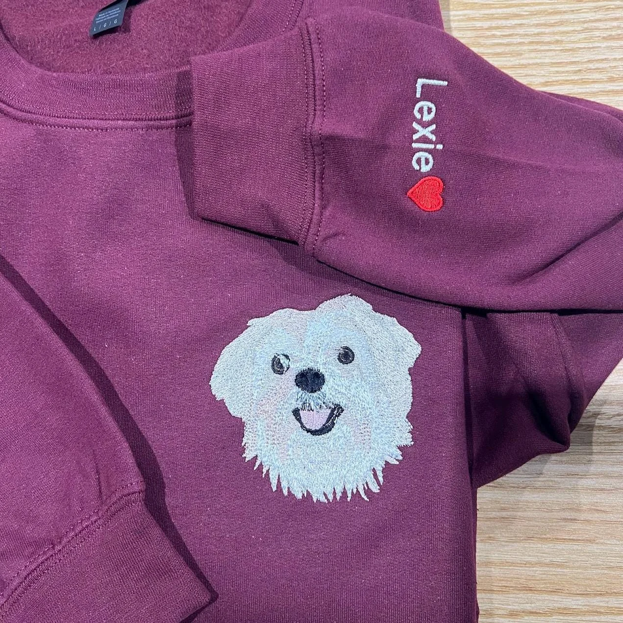 Maroon sweatshirt laid flat with a white dog portrait on the chest and Lexie with a red heart stitched up the sleeve