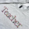 Grey crewneck with floral lettered Teacher embroidery running diagonally down from the collar