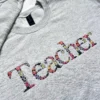 Grey sweatshirt with Teacher embroidered at an angle in letters made of pink, purple and yellow flowers