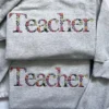 Two grey sweatshirts stacked, each with Teacher spelled in serif letters filled with tiny embroidered flowers