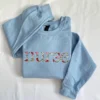 Light blue crewneck laid flat with nurse embroidered across the chest in letters made of colourful flowers