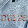 Close-up of nurse in floral embroidered letters with pink roses and white daisies on a light blue sweatshirt