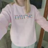 Woman in a light pink crewneck with the word nurse embroidered in multicolour floral letters, mirror view