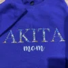 Purple sweatshirt with AKITA spelled in floral embroidered capitals and mom in light blue script below