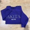 Purple sweatshirt laid on a wooden floor with the floral pet name AKITA and mom across the chest