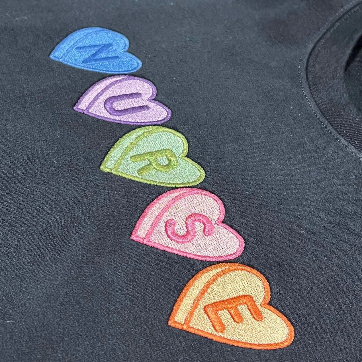 Close-up of five embroidered candy hearts in blue, purple, green, pink and orange, each holding a letter of NURSE