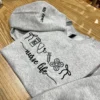 Grey crewneck on a wooden table showing the nurse life icon row and the sleeve name in black thread