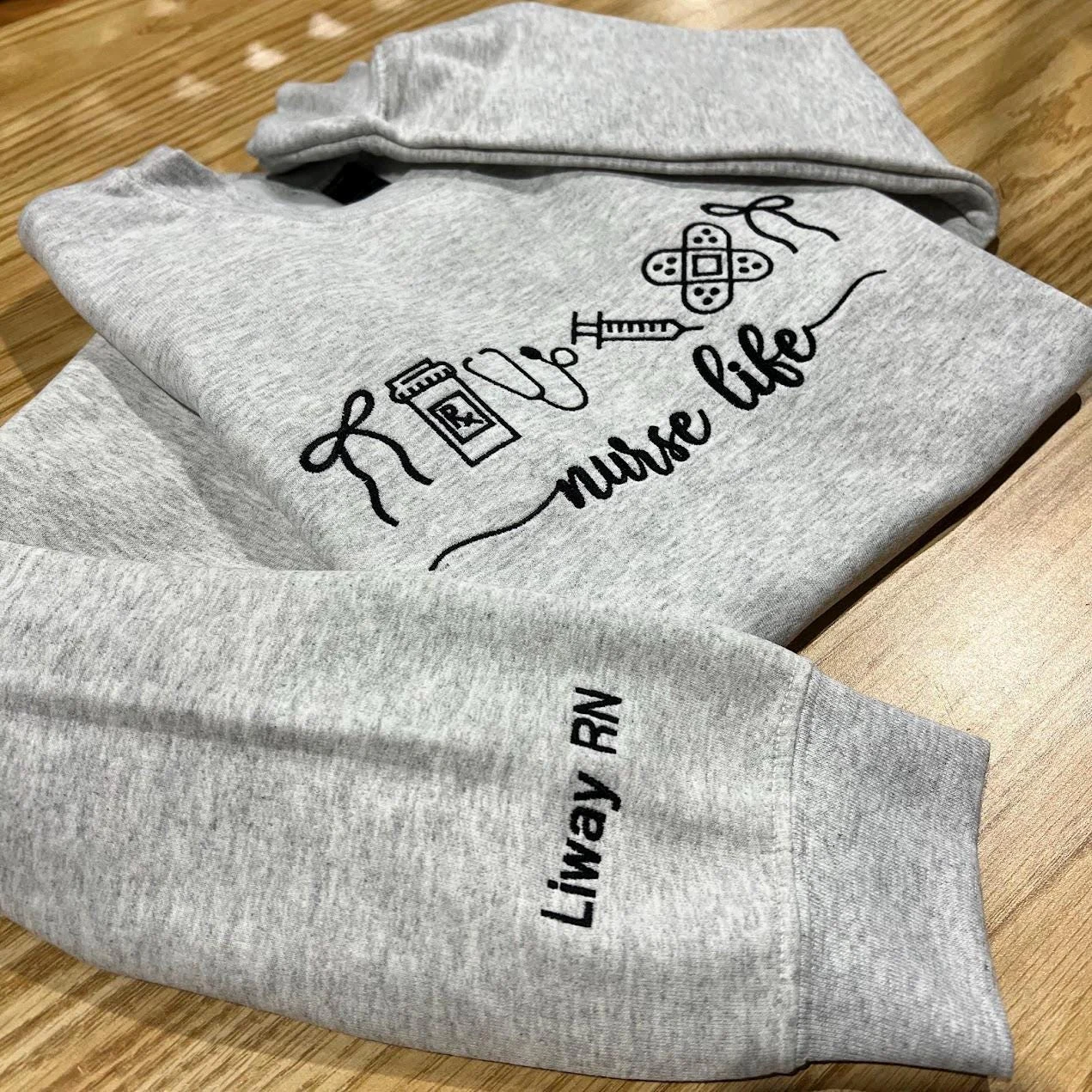 Heather grey nurse life sweatshirt folded on a wooden table with Liway RN embroidered in black on the sleeve