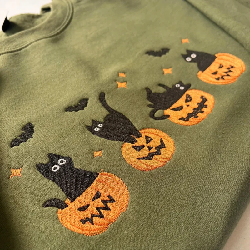Close-up of four black cats on carved jack-o'-lantern pumpkins embroidered on a military green sweatshirt
