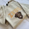 Angled view of a sand sweatshirt with arched ZEUS varsity lettering and a smiling chocolate Labrador portrait