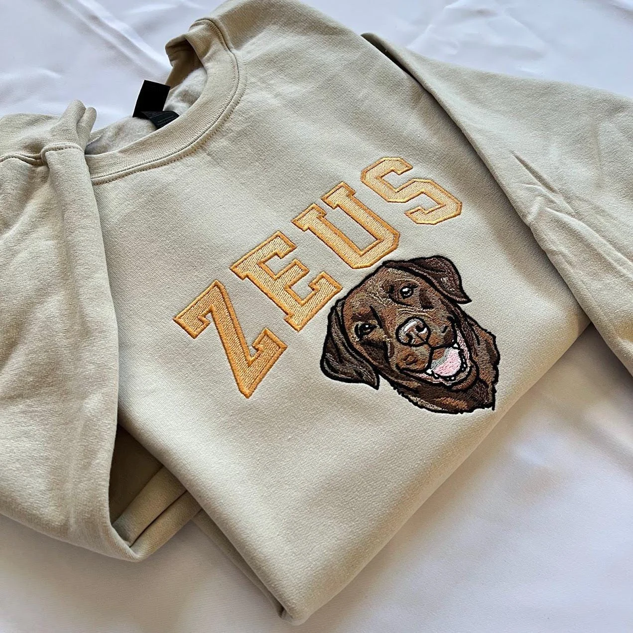 Angled view of a sand sweatshirt with arched ZEUS varsity lettering and a smiling chocolate Labrador portrait