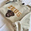 Sand sweatshirt showing a stitched chocolate Labrador with pink tongue beneath tan ZEUS varsity letters