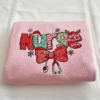 Folded light pink sweatshirt with festive Nurse lettering, a large red bow, stethoscope and white snowflakes