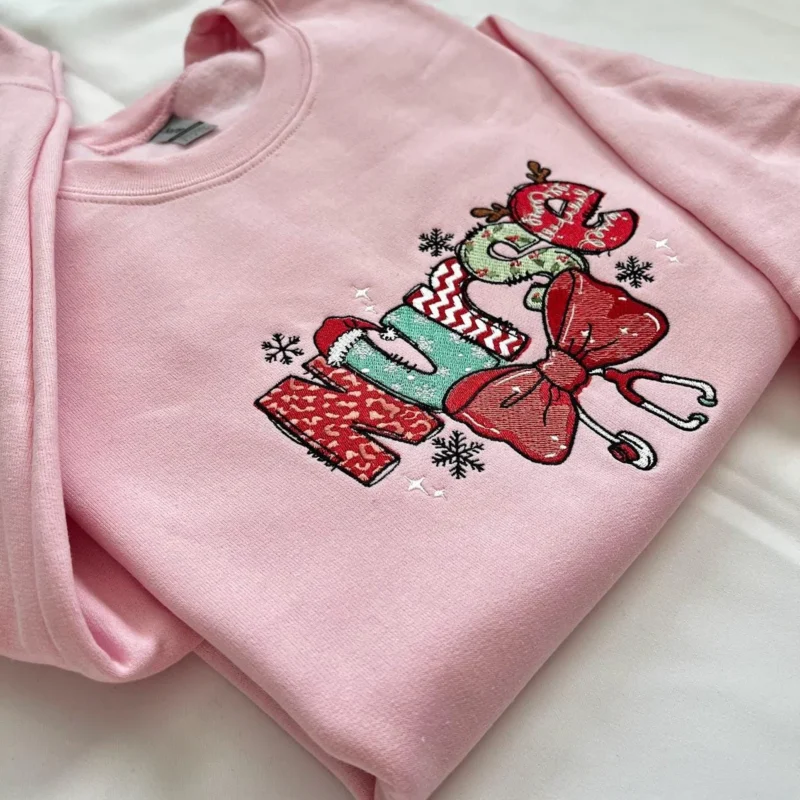 Light pink sweatshirt with Nurse in red and green holiday-pattern letters, a red bow with stethoscope and snowflakes