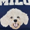Close-up of a cream curly-haired dog face with pink tongue embroidered under white varsity letters on navy