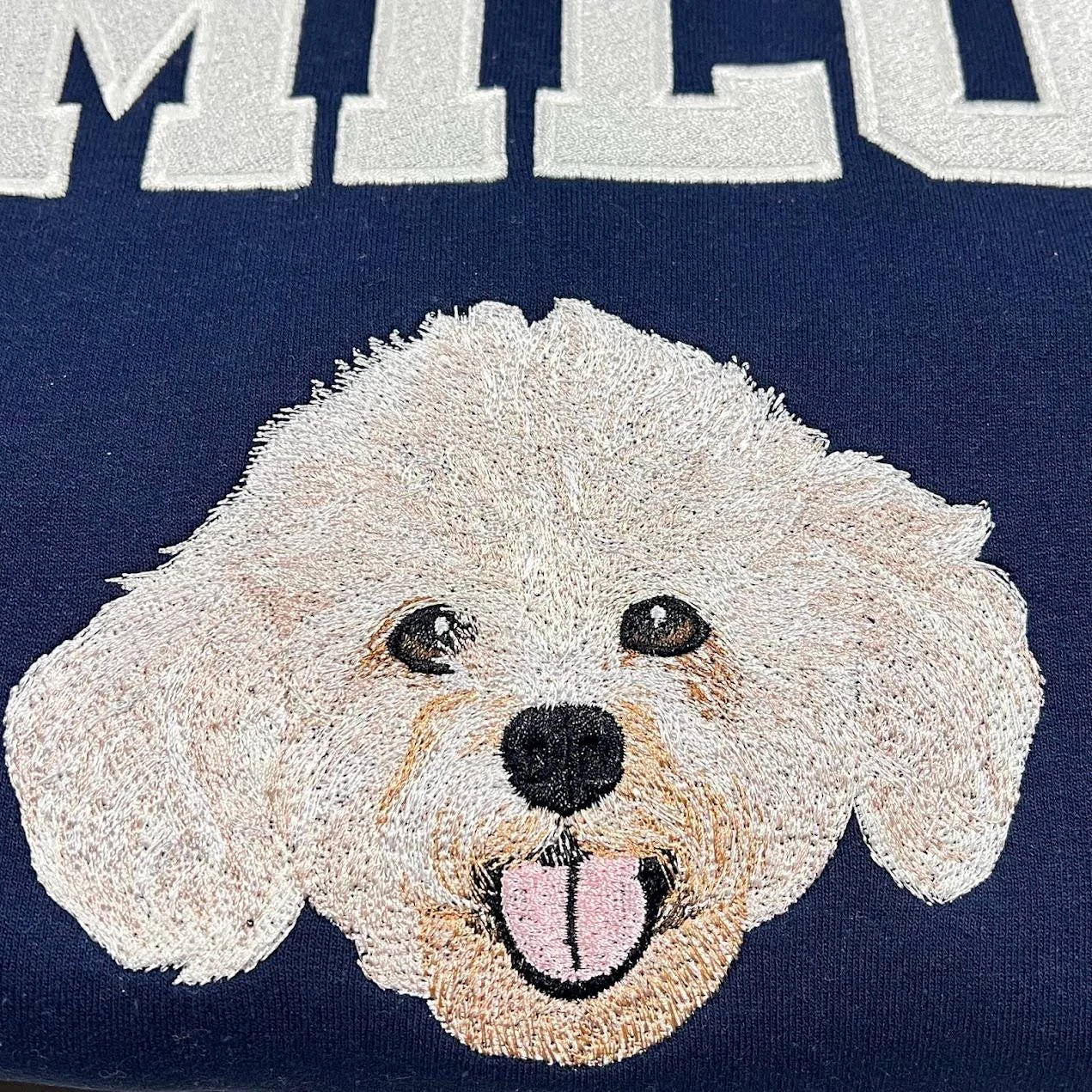 Close-up of a cream curly-haired dog face with pink tongue embroidered under white varsity letters on navy