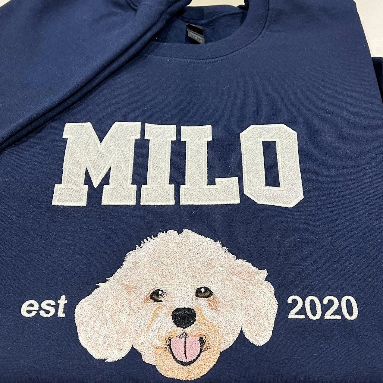 Navy crewneck showing a thread-painted cream curly dog face beneath the name MILO, with est and 2020 either side
