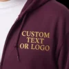 Close-up of a maroon full-zip hoodie with CUSTOM TEXT OR LOGO in gold serif capitals on the left chest