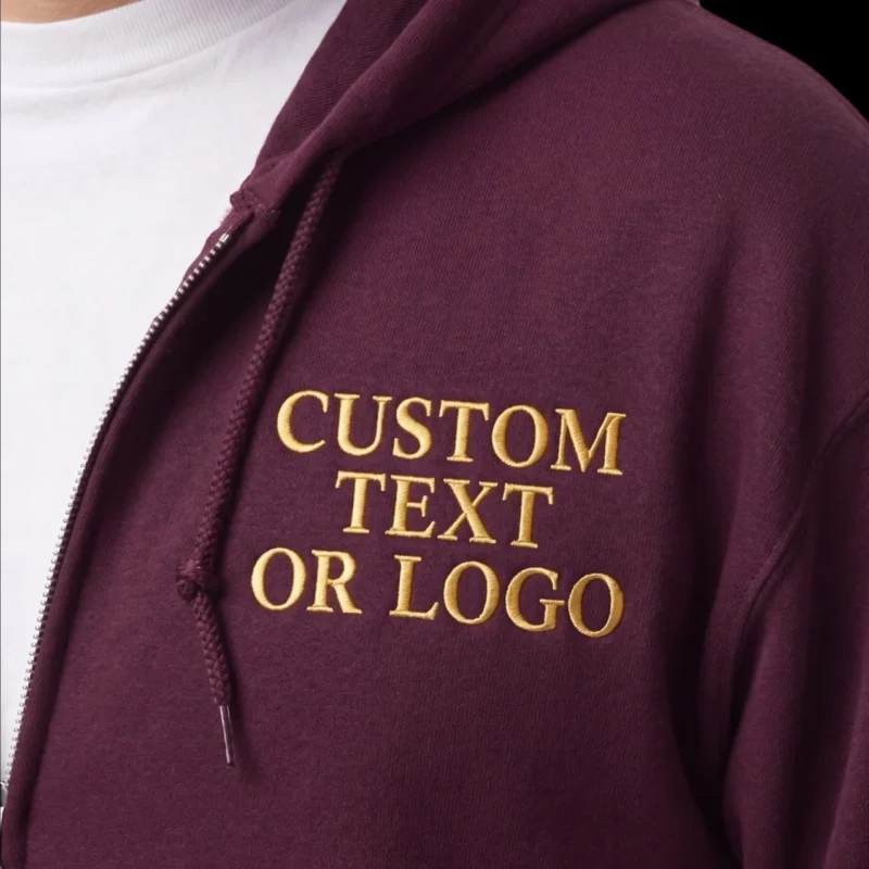 Close-up of a maroon full-zip hoodie with CUSTOM TEXT OR LOGO in gold serif capitals on the left chest