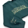 Angled forest green crewneck showing MAMA in cream outlined varsity letters and two names stitched on the cuff