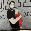 Dad portrait embroidery on a grey sweatshirt, close-up: a bearded dad in black holding a baby in red, EST. 2025