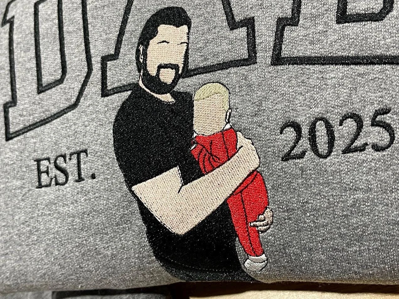 Dad portrait embroidery on a grey sweatshirt, close-up: a bearded dad in black holding a baby in red, EST. 2025