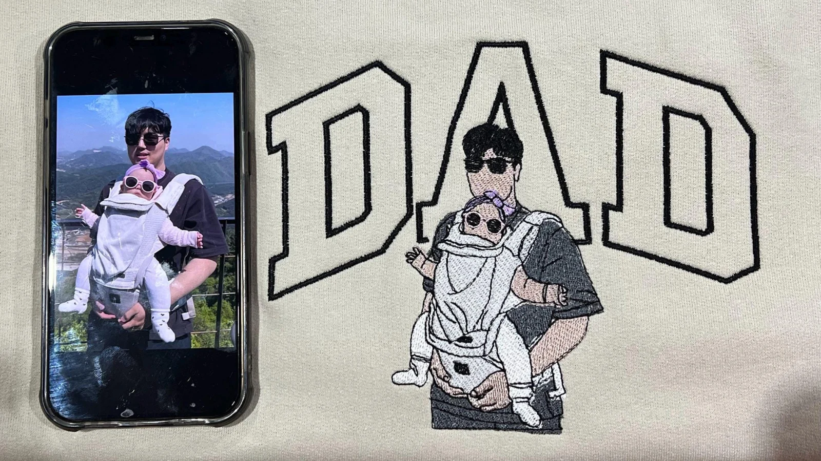Dad portrait embroidery on a cream sweatshirt: black outline DAD and a dad carrying his baby, beside the source photo