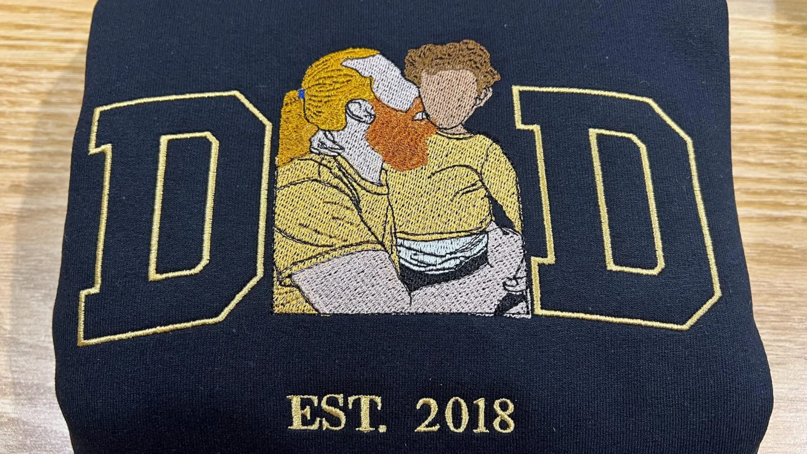 Dad portrait embroidery on a navy sweatshirt: gold outline D and D framing a dad and child in place of the A, EST. 2018