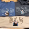 Blue and sand DAD sweatshirts and a charcoal DAD hoodie, each with a stitched dad holding his baby