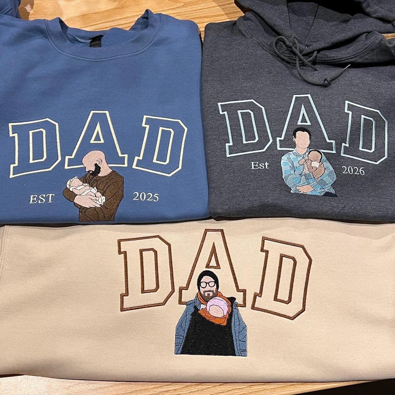Blue and sand DAD sweatshirts and a charcoal DAD hoodie, each with a stitched dad holding his baby