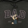 Dark grey hoodie with lavender outline DAD letters and an embroidered dad lifting his baby overhead, EST 2025