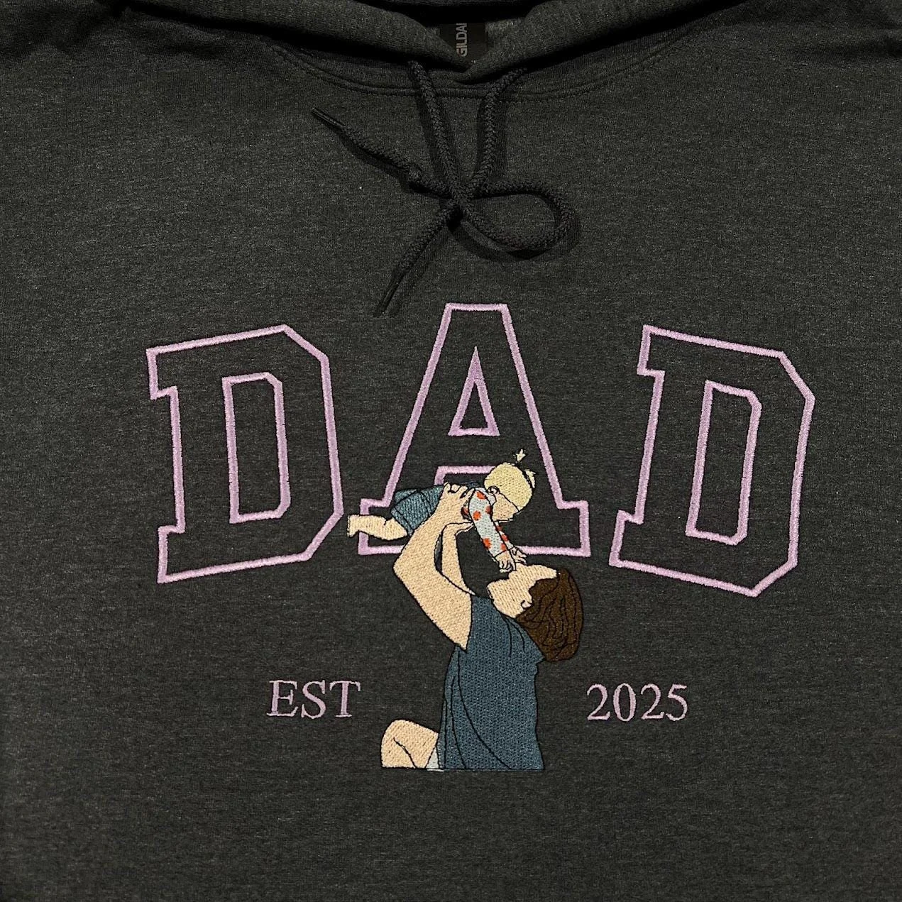 Dark grey hoodie with lavender outline DAD letters and an embroidered dad lifting his baby overhead, EST 2025