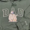 Forest green hoodie with white outline DAD, a dad in a cap holding his baby, and the name Evvy stitched on the sleeve
