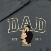 Dad portrait embroidery on a charcoal sweatshirt: cream outline DAD and a dad holding a baby in pink, EST 2025