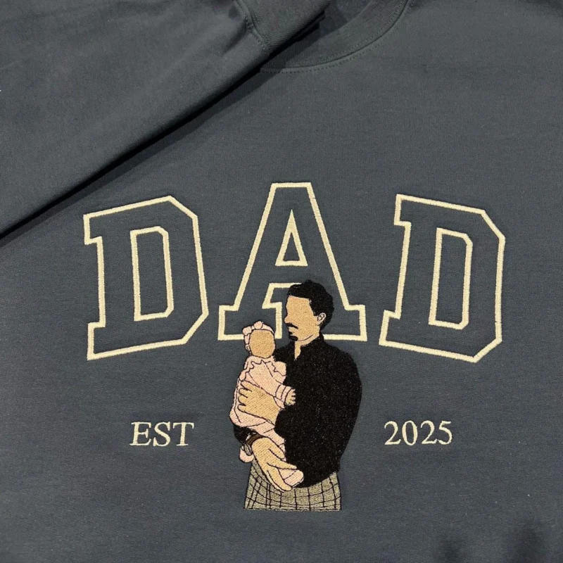 Dad portrait embroidery on a charcoal sweatshirt: cream outline DAD and a dad holding a baby in pink, EST 2025
