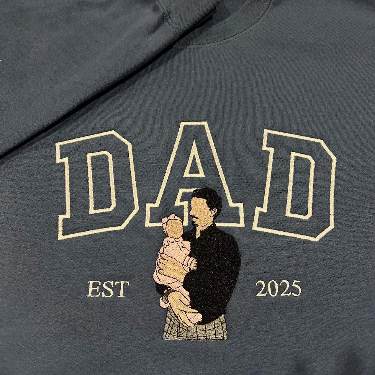 Dad portrait embroidery on a charcoal sweatshirt: cream outline DAD and a dad holding a baby in pink, EST 2025
