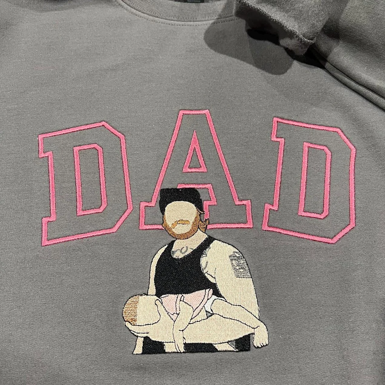 Dad portrait embroidery on a grey sweatshirt: pink outline DAD and a tattooed dad in a cap cradling his baby